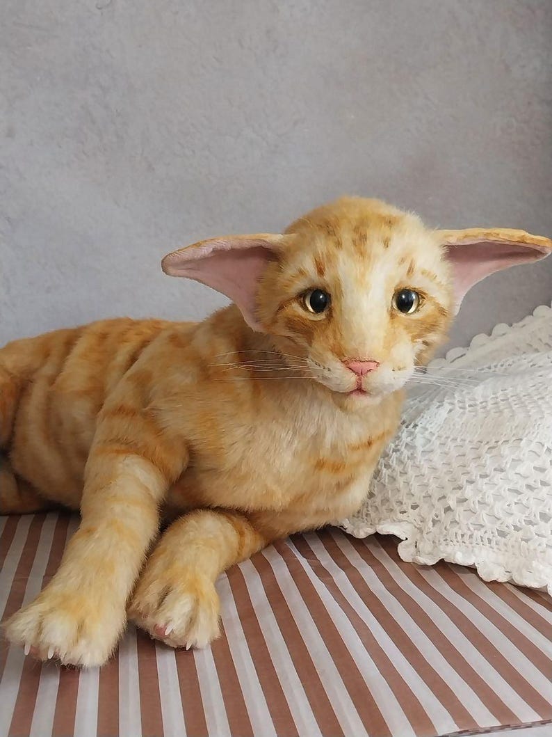 Realistic soft stuffed orange tabby Oriental breed cat toy has yellow taxidermy eyes, pink colored polymer clay paws, white translucent polymer clay claws, and artificial whiskers. The cat  has a green scarf with a diamond pattern. Perfect for collectors, gifts, memorial pet, and home decor.