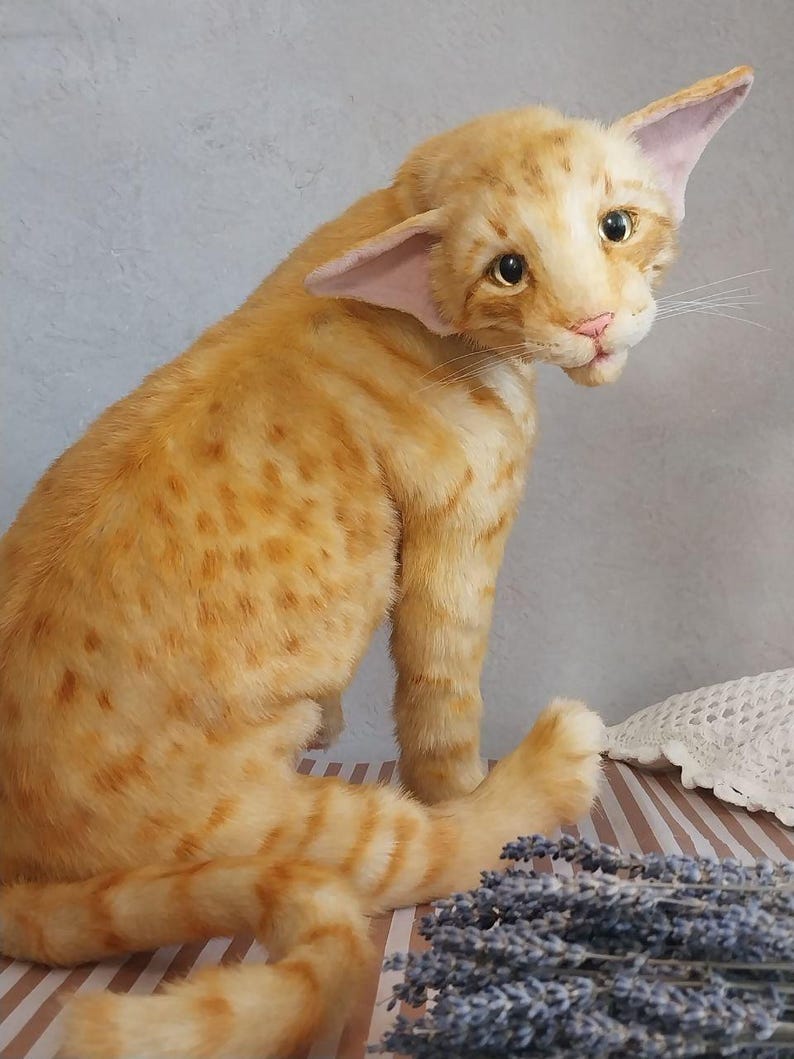Realistic soft stuffed orange tabby Oriental breed cat toy has yellow taxidermy eyes, pink colored polymer clay paws, white translucent polymer clay claws, and artificial whiskers. The cat  has a green scarf with a diamond pattern. Perfect for collectors, gifts, memorial pet, and home decor.