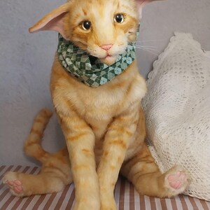 Realistic soft stuffed orange tabby Oriental breed cat toy has yellow taxidermy eyes, pink colored polymer clay paws, white translucent polymer clay claws, and artificial whiskers. The cat  has a green scarf with a diamond pattern. Perfect for collectors, gifts, memorial pet, and home decor.