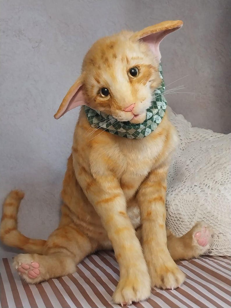 Realistic soft stuffed orange tabby Oriental breed cat toy has yellow taxidermy eyes, pink colored polymer clay paws, white translucent polymer clay claws, and artificial whiskers. The cat  has a green scarf with a diamond pattern. Perfect for collectors, gifts, memorial pet, and home decor.