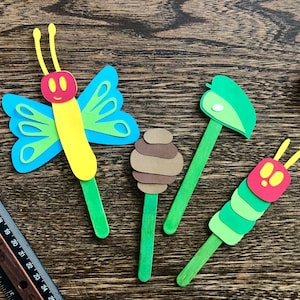May include: Four craft stick puppets depicting characters from the children's book The Very Hungry Caterpillar. The puppets include a green caterpillar, a brown cocoon, a green leaf, and a blue and yellow butterfly.