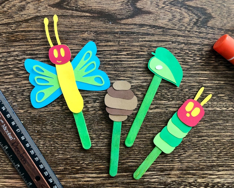 May include: Four craft stick puppets depicting characters from the children's book The Very Hungry Caterpillar. The puppets include a green caterpillar, a brown cocoon, a green leaf, and a blue and yellow butterfly.