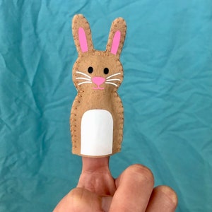 Puede incluir: Un títere de dedo de fieltro marrón con forma de conejo con orejas rosas y vientre blanco. El conejo tiene ojos negros y una nariz rosa.