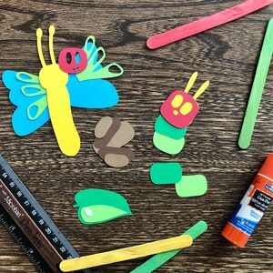 May include: A craft project with paper cutouts of a butterfly, caterpillar, and leaves. The project includes two wooden craft sticks, one green and one yellow, and a bottle of glue.