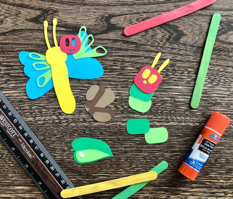 May include: A craft project with paper cutouts of a butterfly, caterpillar, and leaves. The project includes two wooden craft sticks, one green and one yellow, and a bottle of glue.