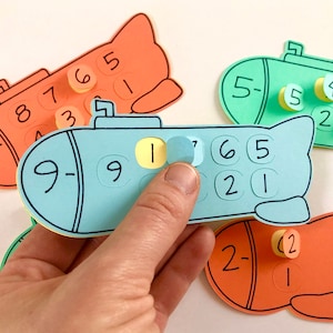 May include: A blue submarine-shaped card with the numbers 9-9, 1, 7, 6, 5, 2, and 1 printed on it. There are yellow and blue circles with numbers on them that can be placed on the card. There are three other cards in the background, two orange and one green, all shaped like submarines with numbers on them.