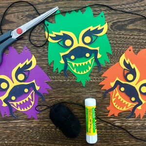 Könnte beinhalten: Drei Papierdrachenmasken in Grün, Lila und Orange mit gelben und schwarzen Details. Die Masken sind mit schwarzem Faden zusammengefügt und es gibt eine Spule schwarzen Fadens, eine Schere und einen Klebestift auf einer Holzoberfläche.