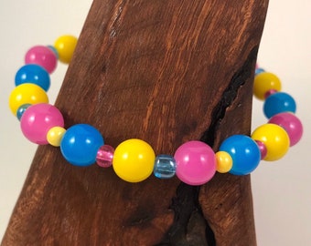 May include: A colorful beaded bracelet with yellow, pink, and blue beads. The bracelet is made with round, smooth beads and is displayed on a wooden surface.