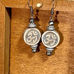 May include: A pair of silver earrings with a gray and white ceramic bead featuring a stylized symbol. The beads are accented with dark gray crystal beads.