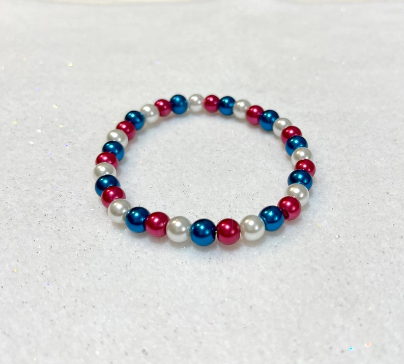 May include: A white, blue, and red beaded bracelet. The beads are arranged in a repeating pattern.