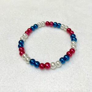 May include: A white, blue, and pink beaded bracelet. The beads are arranged in a repeating pattern.