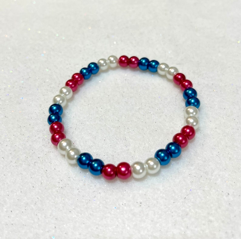 May include: A white, blue, and pink beaded bracelet. The beads are arranged in a repeating pattern.