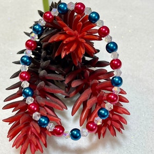 May include: A bracelet with red, blue, and clear beads. The bracelet has alternating red and blue beads, with clear, faceted beads in between. It is displayed on a red and brown floral arrangement, with a white background.