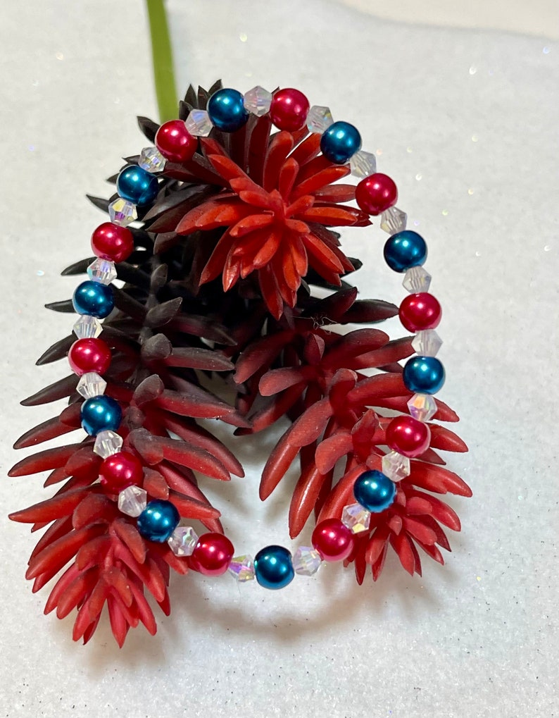 May include: A bracelet with red, blue, and clear beads. The bracelet has alternating red and blue beads, with clear, faceted beads in between. It is displayed on a red and brown floral arrangement, with a white background.