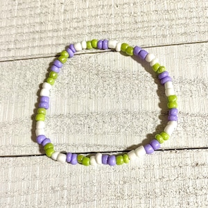 May include: A purple, white, and green beaded bracelet. The bracelet is made of small, round beads that are strung together in a repeating pattern.