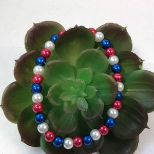 May include: A red, white, and blue beaded bracelet. The bracelet is made of small, round beads and is strung on a clear elastic cord.