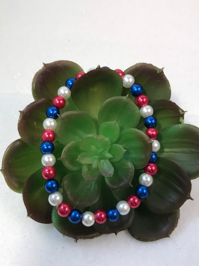 May include: A red, white, and blue beaded bracelet. The bracelet is made of small, round beads and is strung on a clear elastic cord.