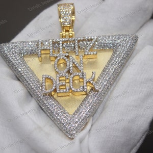 May include: A gold pendant with a triangle shape and the words "HUSTLE ON DECK" set in diamonds. The pendant is on a white surface.