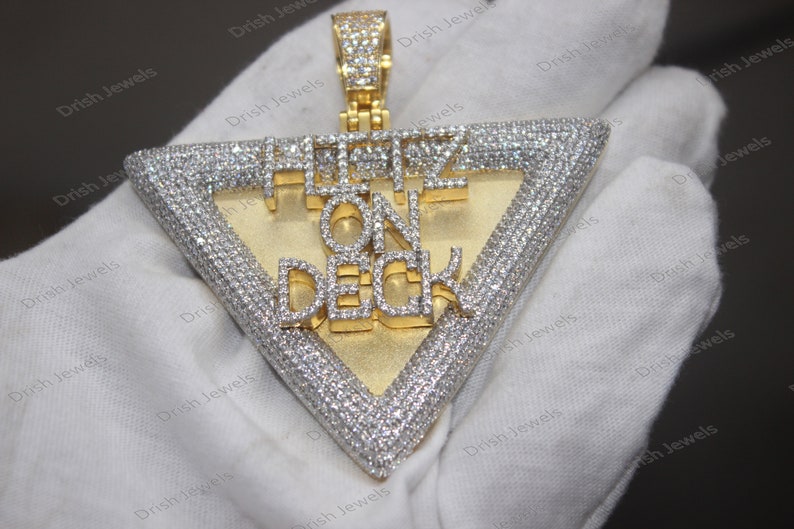May include: A gold pendant with a triangle shape and the words "HUSTLE ON DECK" set in diamonds. The pendant is on a white surface.