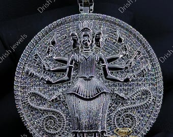 May include: A silver pendant with a detailed design featuring a central figure with multiple arms, surrounded by intricate patterns and sparkling embellishments. The pendant has a rectangular bail and is likely made of a precious metal.