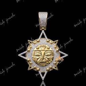 May include: A gold pendant featuring a central sun-like face with stylized features, surrounded by a star-shaped frame encrusted with small white stones. The bail is also paved with white stones, and the entire piece has a polished gold finish.