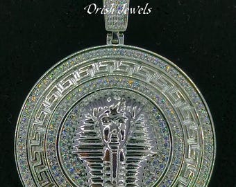 May include: Silver pendant necklace with a round, clear crystal-encrusted design featuring a Pharaoh's head in the center. The pendant is surrounded by a Greek key pattern. The necklace is on a silver rope chain. "Dris Jewels" is written on the pendant.