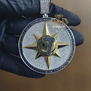 May include: A silver and gold pendant with a large, round, diamond-encrusted design. The center of the pendant features a gold star with a black number 7 inside. The pendant is surrounded by a black diamond border.