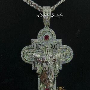May include: A silver and white diamond encrusted pendant featuring a depiction of Jesus Christ on a cross. The pendant has red gemstones and the inscription "IC XC".