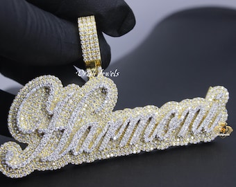 Puede incluir: Un colgante de oro con el nombre "Jamaica" escrito en letras grandes y en negrita. Las letras están cubiertas de piedras de circonita cúbica transparentes y brillantes.