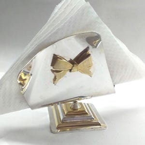 May include: Silver napkin holder with a gold butterfly design. The holder has a square base and a triangular top.