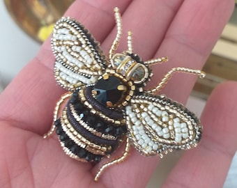 May include: A beaded bee brooch with black and white wings, a black body, and gold accents. The bee has a black gem for a head.