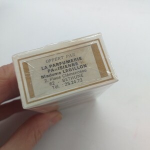 May include: A white box with a brown border and gold accents. The box has text that reads "OFFERT PAR LA PARFUMERIE PARISIENNE Madame LEGILLON 2, Place Cl&eacute;menceau 62 - BETHUNE T&eacute;l.: 25.24.72"