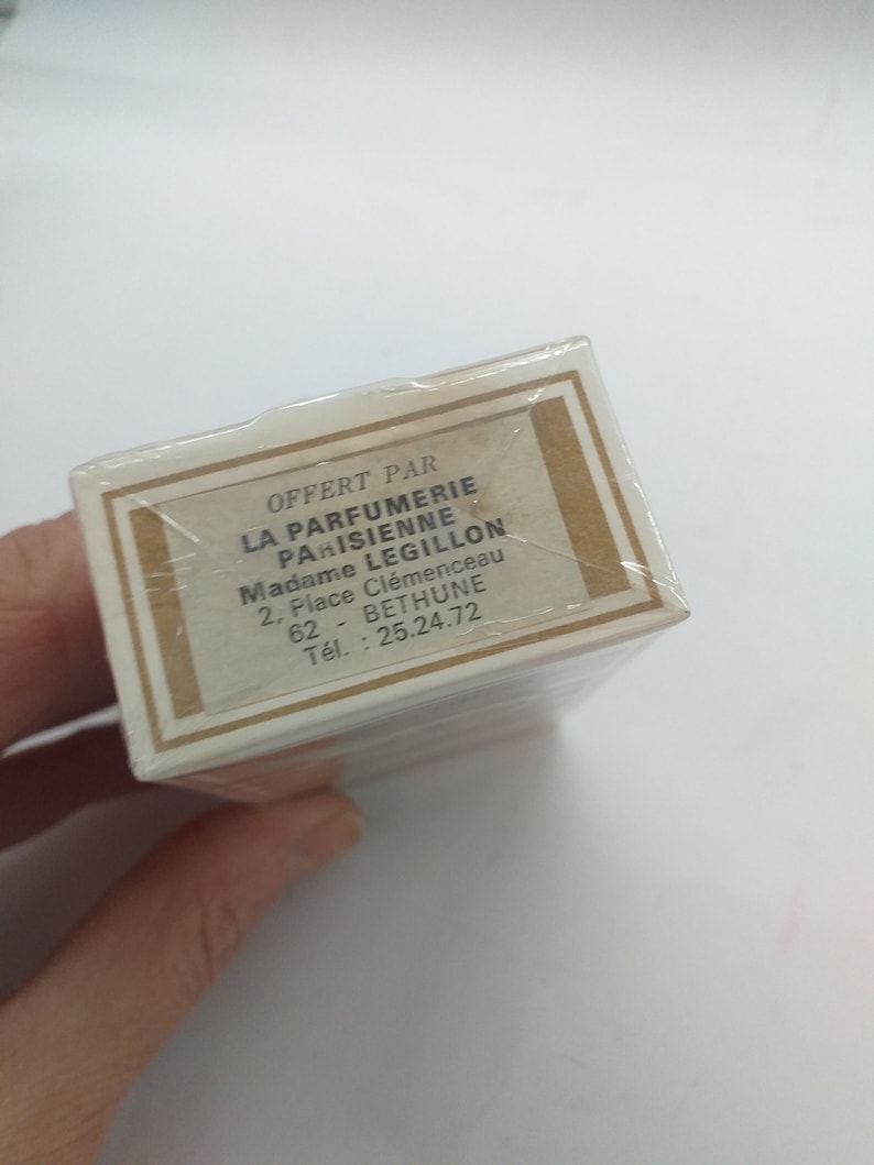 May include: A white box with a brown border and gold accents. The box has text that reads "OFFERT PAR LA PARFUMERIE PARISIENNE Madame LEGILLON 2, Place Cl&eacute;menceau 62 - BETHUNE T&eacute;l.: 25.24.72"