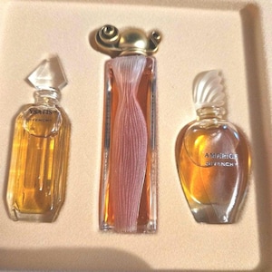 May include: A set of three Givenchy perfume bottles in a beige box. The bottles are filled with amber-coloured liquid. The bottles have different shapes and designs, including a dress-shaped bottle and bottles with the text "YSATIS" and "AMARIGE GIVENCHY".