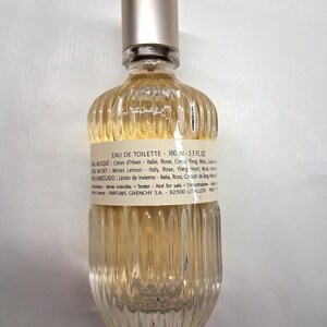 May include: A clear glass Eau de Toilette bottle with a silver cap. The bottle has a ribbed design and contains a light yellow liquid. The label includes text about the fragrance's ingredients and volume, which is 3.3 fl oz.