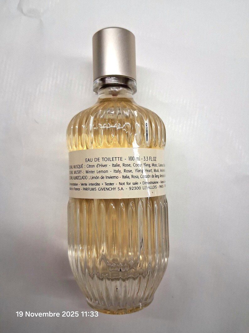 May include: A clear glass Eau de Toilette bottle with a silver cap. The bottle has a ribbed design and contains a light yellow liquid. The label includes text about the fragrance's ingredients and volume, which is 3.3 fl oz.