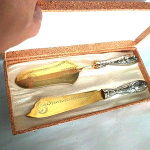 May include: A gold-toned cake server and knife set with ornate silver-toned handles, presented in a rectangular box with a textured brown exterior and a white satin-lined interior. Both utensils feature delicate engraved floral patterns.