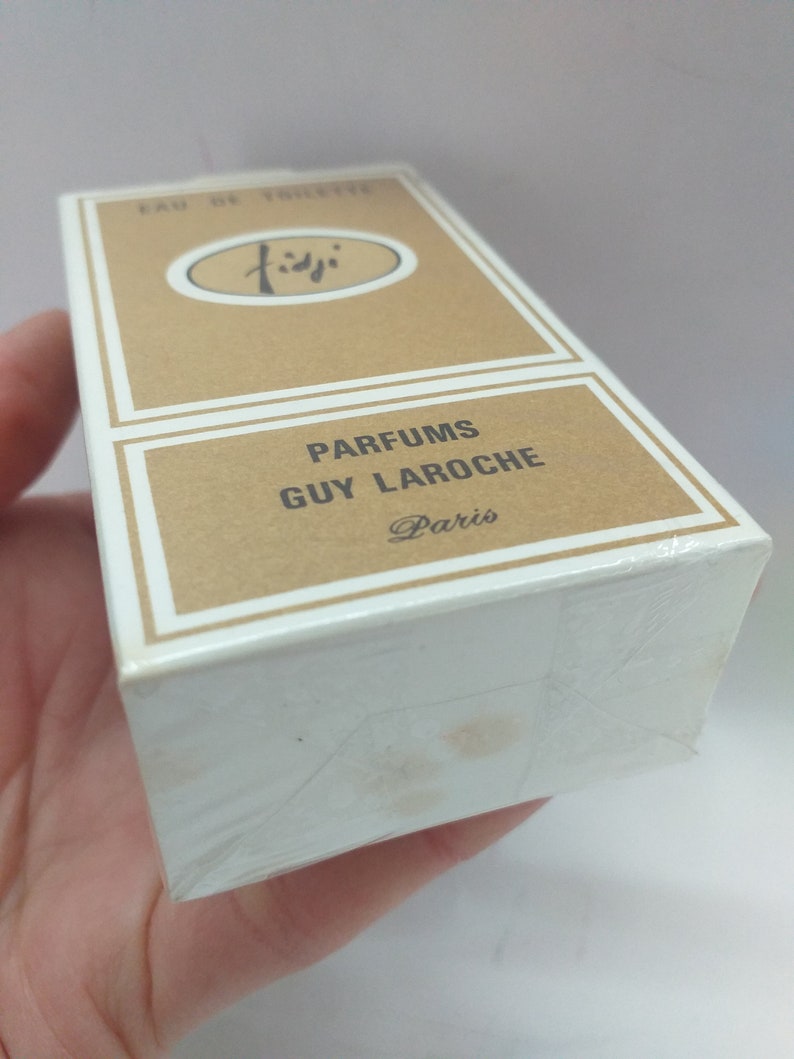 May include: A brown cardboard box with white trim, featuring the text "PARFUMS GUY LAROCHE Paris" and a logo with the word "Fidji" in a circle. The box is wrapped in clear plastic.