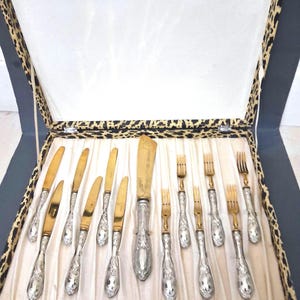 May include: A set of vintage silverware, including knives and forks, presented in a leopard print lined box. The silverware features ornate silver handles and gold-toned blades and tines. The box is lined with a cream-colored fabric.