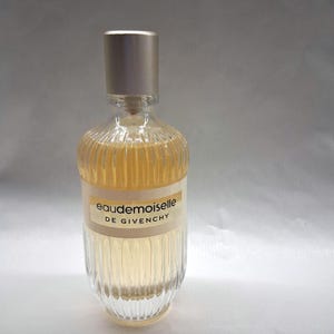 May include: A clear glass perfume bottle with a gold-colored cap. The bottle has a ribbed design and contains a light amber liquid. The label reads "Eaudemoiselle DE GIVENCHY".