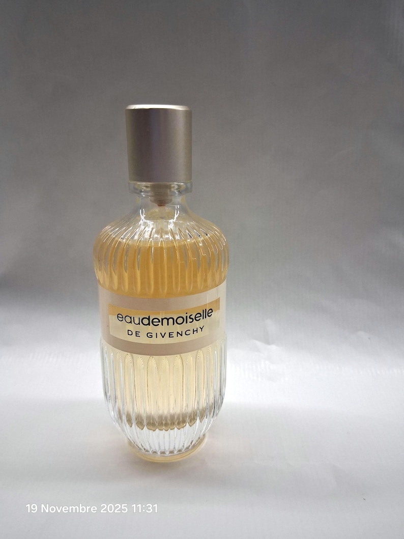 May include: A clear glass perfume bottle with a gold-colored cap. The bottle has a ribbed design and contains a light amber liquid. The label reads "Eaudemoiselle DE GIVENCHY".