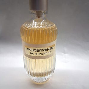 May include: A clear glass perfume bottle with a silver cap. The bottle has vertical ridges and contains a light amber liquid. The label reads "eaudemoiselle DE GIVENCHY".