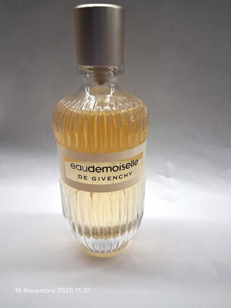 May include: A clear glass perfume bottle with a silver cap. The bottle has vertical ridges and contains a light amber liquid. The label reads "eaudemoiselle DE GIVENCHY".