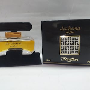 May include: A perfume bottle and box set. The perfume bottle is clear with a yellow liquid inside, a black cap, and the words "detchema parfum" in gold. The black box has gold text and a geometric design, with "Revillon PARIS" and "1/2 fl.oz."