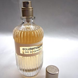 May include: A clear glass perfume bottle with a gold-colored cap and a beige label that reads "Eaudemoiselle DE GIVENCHY." The bottle has a ribbed design and contains a light amber liquid. The cap is off to the side.