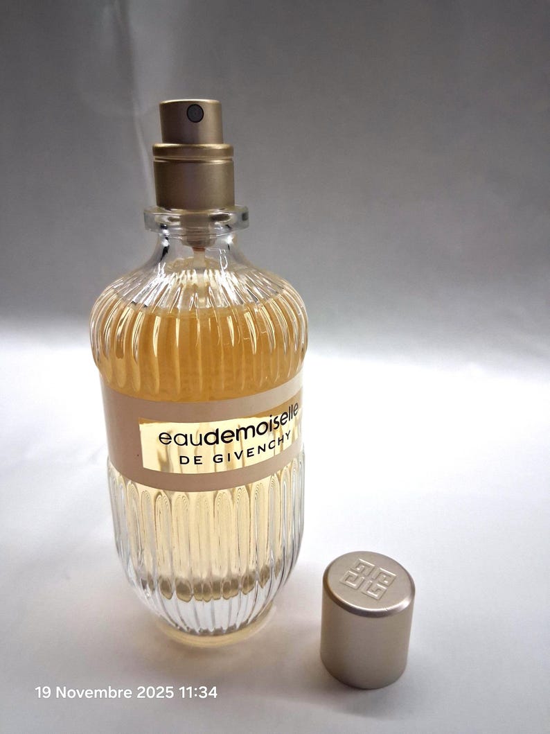 May include: A clear glass perfume bottle with a gold-colored cap and a beige label that reads "Eaudemoiselle DE GIVENCHY." The bottle has a ribbed design and contains a light amber liquid. The cap is off to the side.