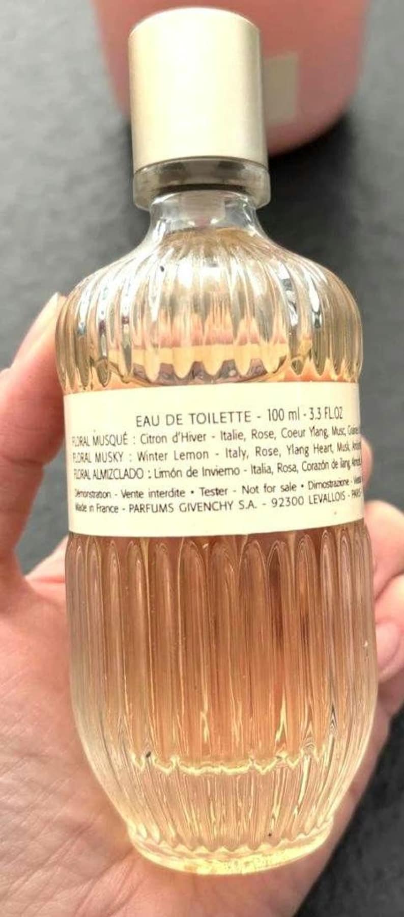 May include: A clear glass bottle of Eau de Toilette, 100 ml (3.3 fl oz), with a silver cap. The bottle has a ribbed design and contains a light amber liquid. Text on the label includes fragrance notes and the brand name.