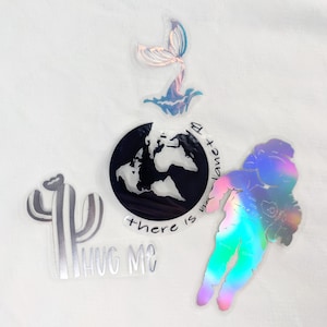 Könnte beinhalten: Ein Set mit fünf holografischen Aufklebern, die eine Meerjungfrauenflosse, einen Kaktus, einen Astronauten, einen Globus mit dem Text "There is no planet B" und einen Kaktus mit dem Text "Hug Me" zeigen.