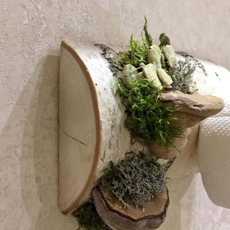 Puede incluir: Un porta rollo de papel higi&eacute;nico de madera de abedul con un dise&ntilde;o r&uacute;stico. El soporte presenta detalles de musgo, hongos y l&iacute;quenes.