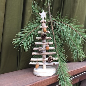 Może przedstawiać: Małe, białe, drewniane drzewko bożonarodzeniowe ozdobione żołędziami i szyszkami. Drzewko stoi na drewnianej powierzchni.